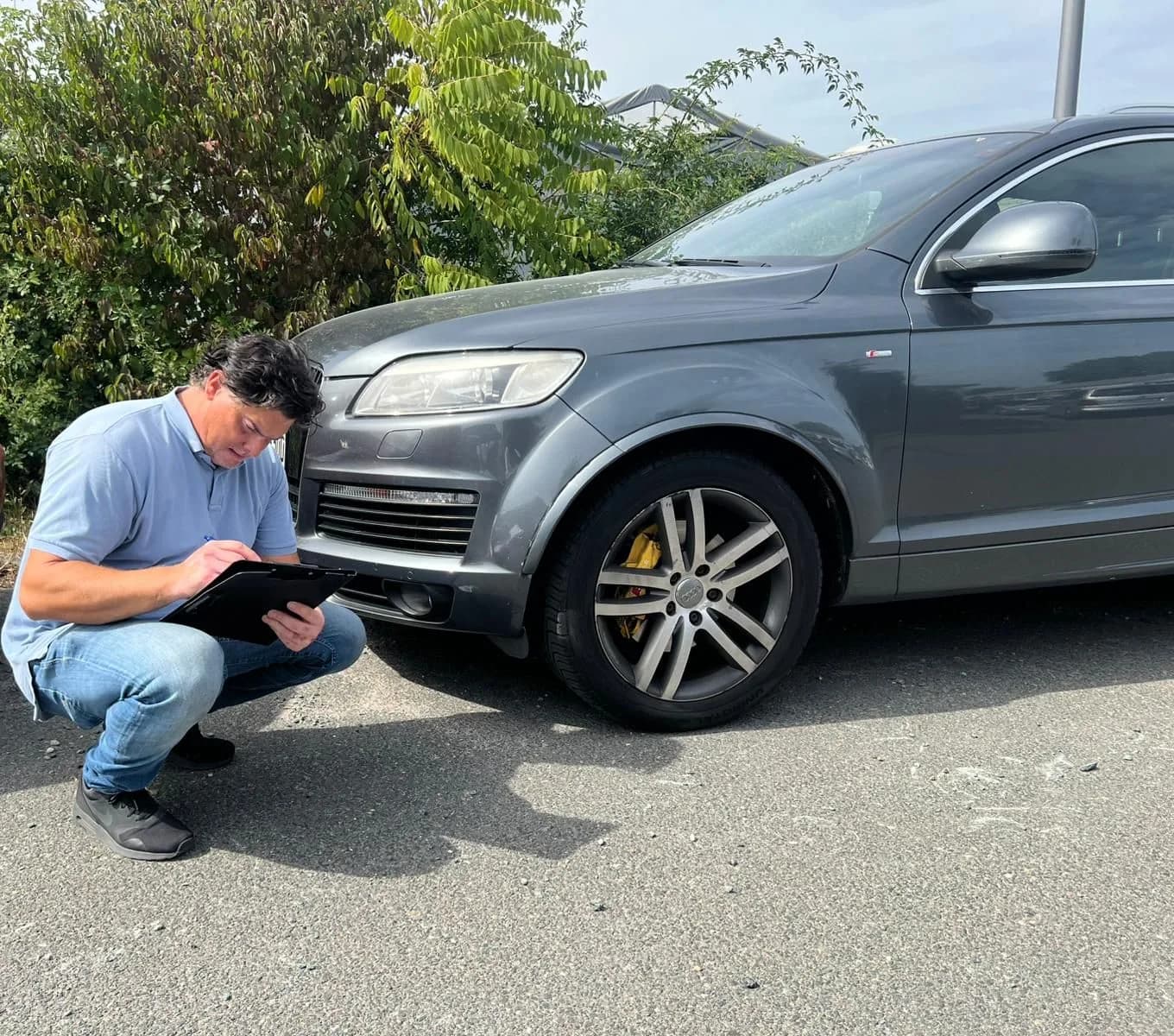Ferdinand Wolf hockt mit Klemmbrett vor der Front eines dunkelgrauen SUV